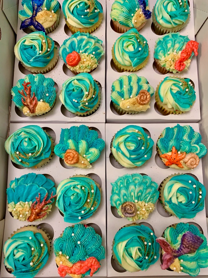 Boxes of teal ocean-themed cupcakes with seashells, coral, and mermaid tails